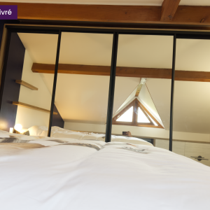 Vue en contre-plongée d'une chambre sous combles avec grand placard à portes miroir, poutres apparentes et mur bleu nuit, reflétant la lumière naturelle de la lucarne.