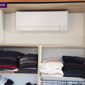 Gros plan sur l’unité de climatisation intégrée dans la niche supérieure du placard, entourée de vêtements pliés sur étagères ouvertes.