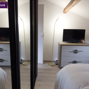 Vue du coin TV dans la chambre, avec une commode en bois clair à deux tiroirs, miroir et lampe dorée design.